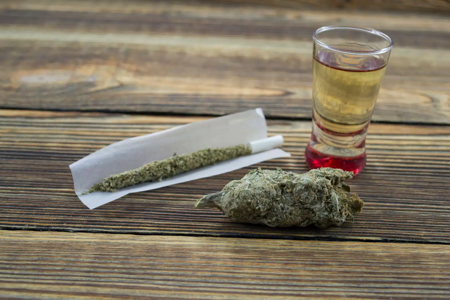An unrolled joint, marijuana bud, and a shot glass are on a wooden tabletop - Is Alcohol Or Cannabis Worse For Your Body
