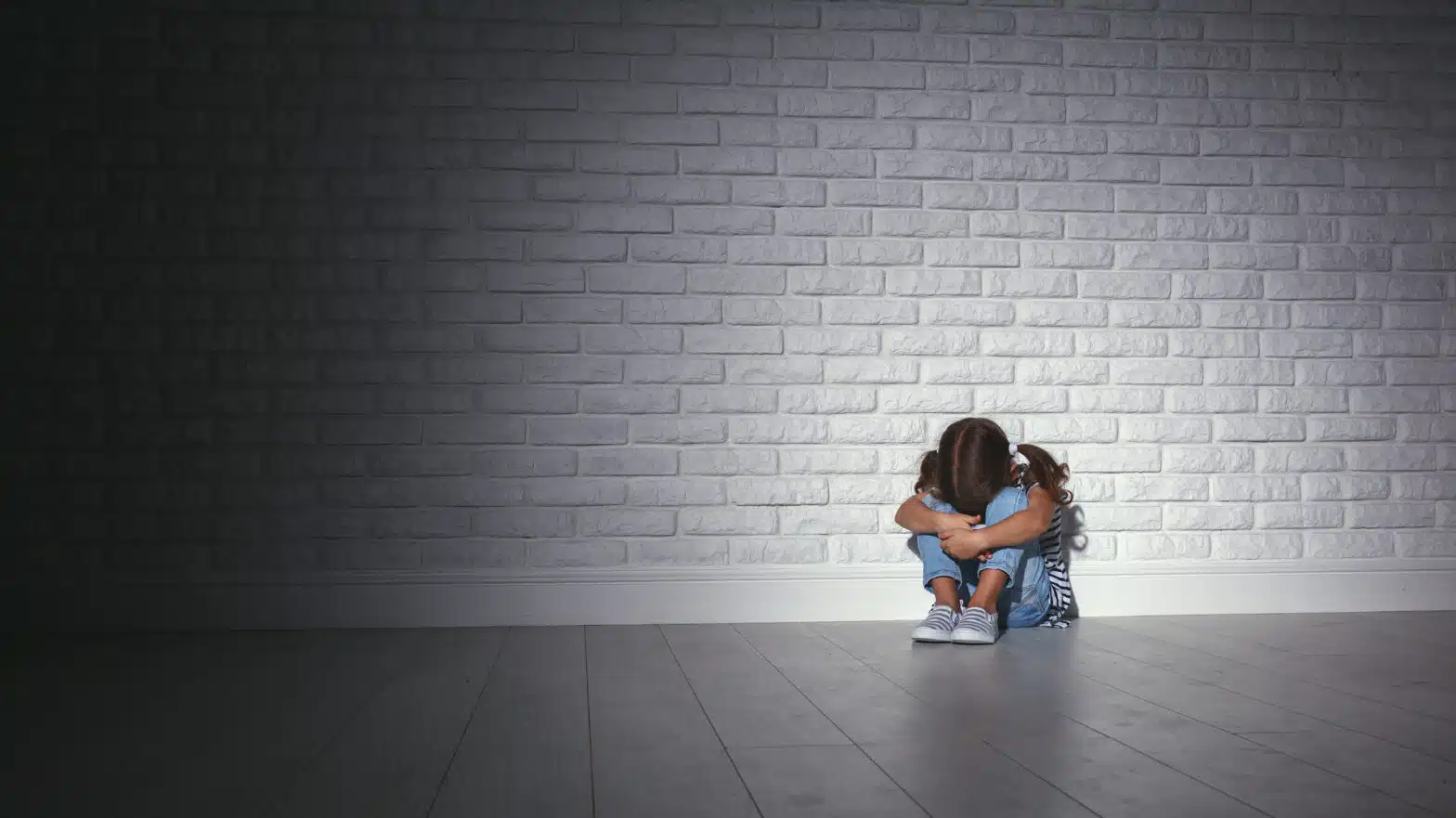A young girl sits curled up in front of a brick wall - The Effects Of Parental Substance Abuse On Children