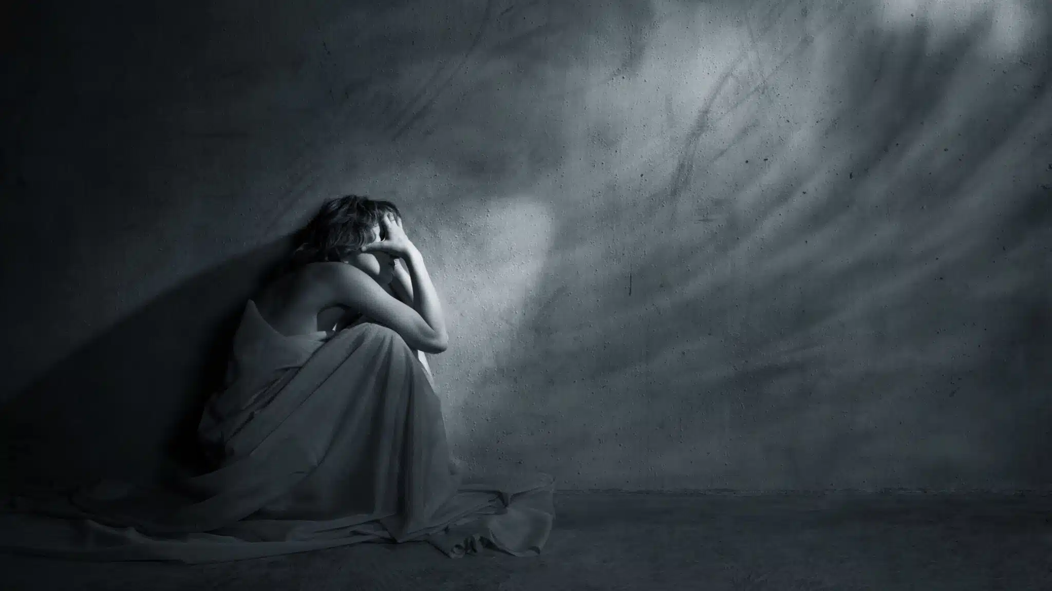 A woman sits alone on the floor in a dark room - Suicide Risks & Prevention In Early Recovery